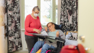 Avustaja ja henkilö pyörätuolissa katsovat paperia. Avustaja ja henkilö pyörätuolissa katsovat paperia.